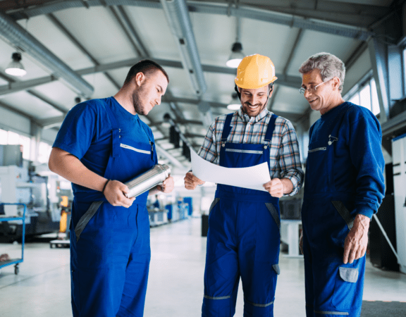 Three men in blue overalls and hard hats looking at papers.