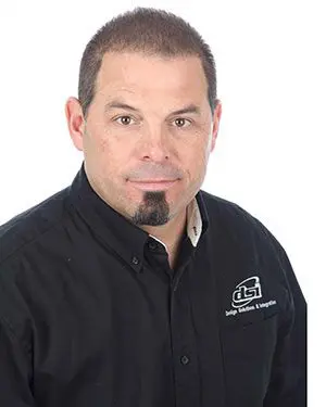 A man in black shirt and beard standing next to white wall.