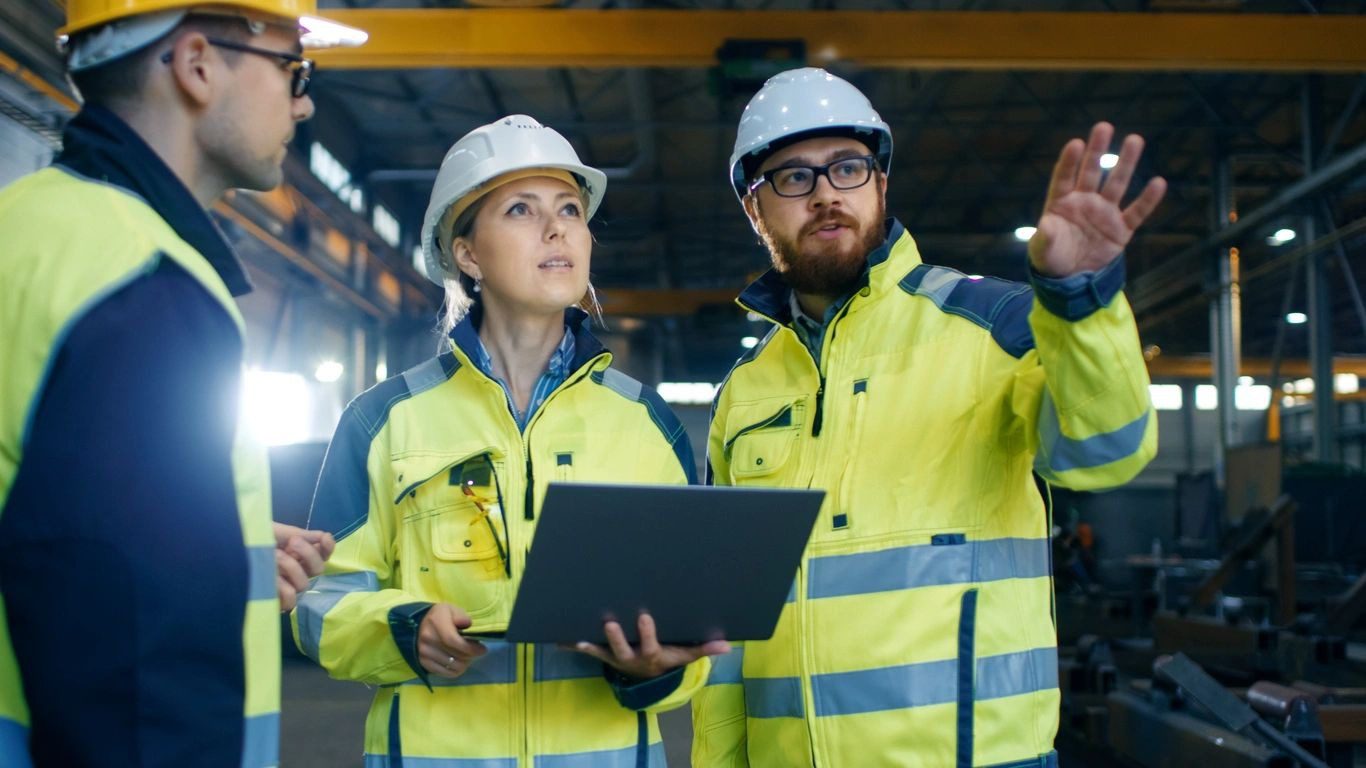 Two people in yellow jackets and hard hats holding a laptop.